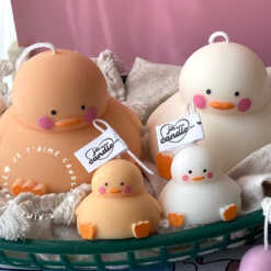 A close-up of four handcrafted duck-shaped candles in soft yellow tones with orange beaks and feet, and pink blushed cheeks, ideal for cute gifts or decorative accents. The candles are labeled with 'Je T’aime Candle' tags and are placed beside a dried floral arrangement, creating a soft, aesthetic backdrop. These adorable soy wax candles are perfect for baby showers, birthday favors, Easter gifts, or aesthetic room decor.