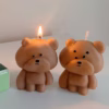 Three bear-shaped candles are placed on a white surface. Two of the candles are lit, with flames on top of their heads. The candles have round heads, small ears, and simple facial features. One candle is positioned on a green-tiled block, while the other two stand directly on the surface.