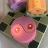 A pink yin-yang candle with two flames sits on a green and white tiled surface. A white gift box with a "Thank you" ribbon is in the background. Cozy and elegant.