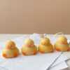 A close-up of four handcrafted duck-shaped candles in soft yellow tones with orange beaks and feet, and pink blushed cheeks, ideal for cute gifts or decorative accents. Three of the duck candles are packaged in clear plastic gift boxes tied with pastel-colored ribbon bows in blue, lavender, and peach, while one candle is displayed unboxed on a layered stack of white papers for a natural, cozy setting. The candles are labeled with 'Je T’aime Candle' tags and are placed beside a dried floral arrangement, creating a soft, aesthetic backdrop. These adorable soy wax candles are perfect for baby showers, birthday favors, Easter gifts, or aesthetic room decor.
