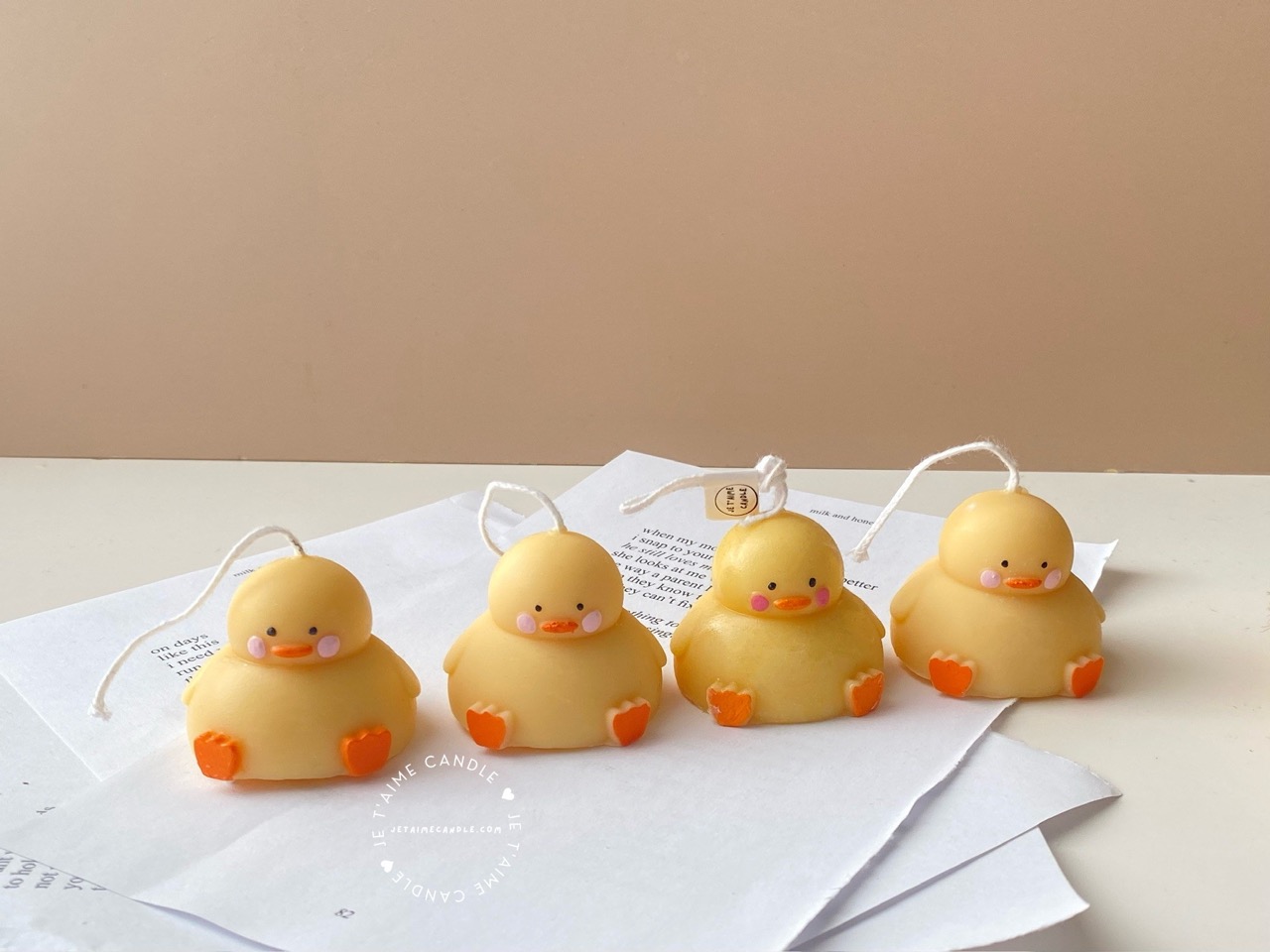 A close-up of four handcrafted duck-shaped candles in soft yellow tones with orange beaks and feet, and pink blushed cheeks, ideal for cute gifts or decorative accents. Three of the duck candles are packaged in clear plastic gift boxes tied with pastel-colored ribbon bows in blue, lavender, and peach, while one candle is displayed unboxed on a layered stack of white papers for a natural, cozy setting. The candles are labeled with 'Je T’aime Candle' tags and are placed beside a dried floral arrangement, creating a soft, aesthetic backdrop. These adorable soy wax candles are perfect for baby showers, birthday favors, Easter gifts, or aesthetic room decor.