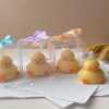 A close-up of four handcrafted duck-shaped candles in soft yellow tones with orange beaks and feet, and pink blushed cheeks, ideal for cute gifts or decorative accents. Three of the duck candles are packaged in clear plastic gift boxes tied with pastel-colored ribbon bows in blue, lavender, and peach, while one candle is displayed unboxed on a layered stack of white papers for a natural, cozy setting. The candles are labeled with 'Je T’aime Candle' tags and are placed beside a dried floral arrangement, creating a soft, aesthetic backdrop. These adorable soy wax candles are perfect for baby showers, birthday favors, Easter gifts, or aesthetic room decor.