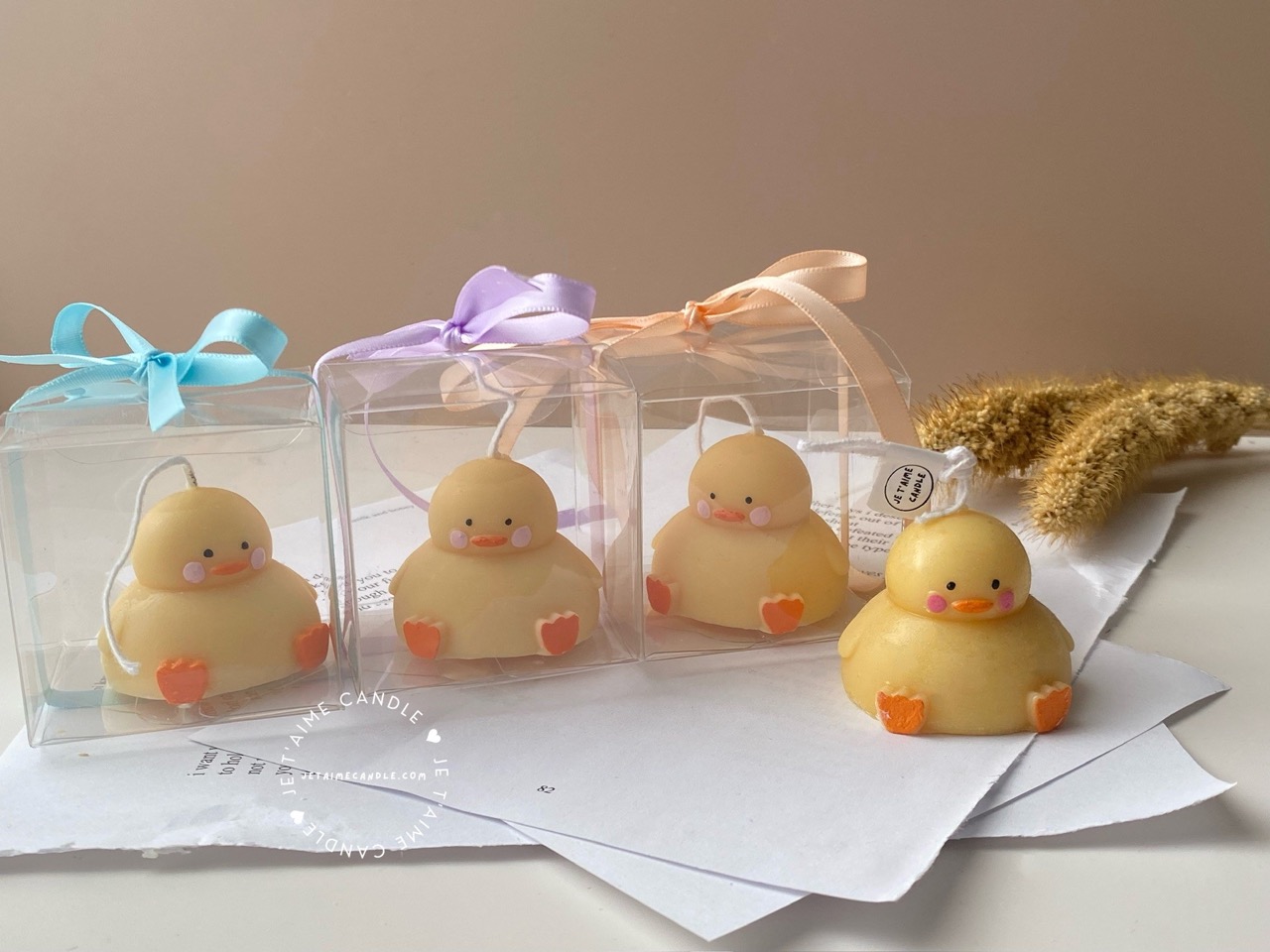 A close-up of four handcrafted duck-shaped candles in soft yellow tones with orange beaks and feet, and pink blushed cheeks, ideal for cute gifts or decorative accents. Three of the duck candles are packaged in clear plastic gift boxes tied with pastel-colored ribbon bows in blue, lavender, and peach, while one candle is displayed unboxed on a layered stack of white papers for a natural, cozy setting. The candles are labeled with 'Je T’aime Candle' tags and are placed beside a dried floral arrangement, creating a soft, aesthetic backdrop. These adorable soy wax candles are perfect for baby showers, birthday favors, Easter gifts, or aesthetic room decor.