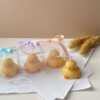 A close-up of four handcrafted duck-shaped candles in soft yellow tones with orange beaks and feet, and pink blushed cheeks, ideal for cute gifts or decorative accents. Three of the duck candles are packaged in clear plastic gift boxes tied with pastel-colored ribbon bows in blue, lavender, and peach, while one candle is displayed unboxed on a layered stack of white papers for a natural, cozy setting. The candles are labeled with 'Je T’aime Candle' tags and are placed beside a dried floral arrangement, creating a soft, aesthetic backdrop. These adorable soy wax candles are perfect for baby showers, birthday favors, Easter gifts, or aesthetic room decor.
