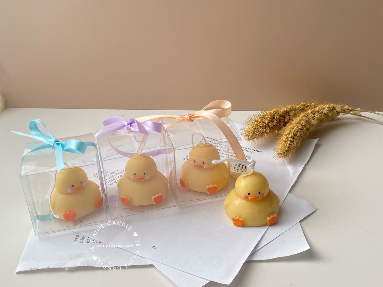 A close-up of four handcrafted duck-shaped candles in soft yellow tones with orange beaks and feet, and pink blushed cheeks, ideal for cute gifts or decorative accents. Three of the duck candles are packaged in clear plastic gift boxes tied with pastel-colored ribbon bows in blue, lavender, and peach, while one candle is displayed unboxed on a layered stack of white papers for a natural, cozy setting. The candles are labeled with 'Je T’aime Candle' tags and are placed beside a dried floral arrangement, creating a soft, aesthetic backdrop. These adorable soy wax candles are perfect for baby showers, birthday favors, Easter gifts, or aesthetic room decor.