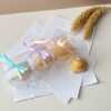 A close-up of four handcrafted duck-shaped candles in soft yellow tones with orange beaks and feet, and pink blushed cheeks, ideal for cute gifts or decorative accents. Three of the duck candles are packaged in clear plastic gift boxes tied with pastel-colored ribbon bows in blue, lavender, and peach, while one candle is displayed unboxed on a layered stack of white papers for a natural, cozy setting. The candles are labeled with 'Je T’aime Candle' tags and are placed beside a dried floral arrangement, creating a soft, aesthetic backdrop. These adorable soy wax candles are perfect for baby showers, birthday favors, Easter gifts, or aesthetic room decor.