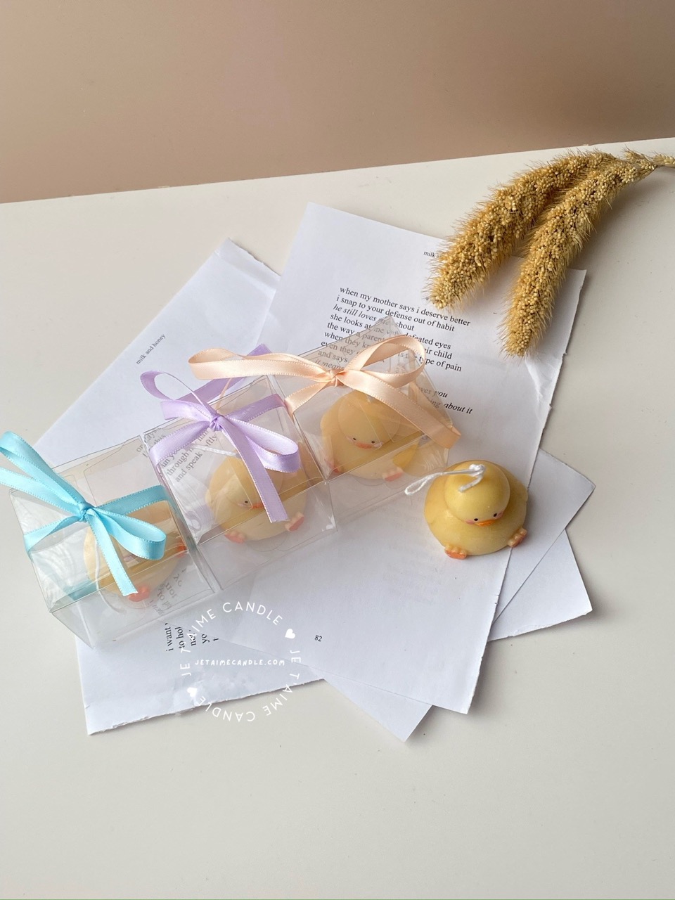 A close-up of four handcrafted duck-shaped candles in soft yellow tones with orange beaks and feet, and pink blushed cheeks, ideal for cute gifts or decorative accents. Three of the duck candles are packaged in clear plastic gift boxes tied with pastel-colored ribbon bows in blue, lavender, and peach, while one candle is displayed unboxed on a layered stack of white papers for a natural, cozy setting. The candles are labeled with 'Je T’aime Candle' tags and are placed beside a dried floral arrangement, creating a soft, aesthetic backdrop. These adorable soy wax candles are perfect for baby showers, birthday favors, Easter gifts, or aesthetic room decor.