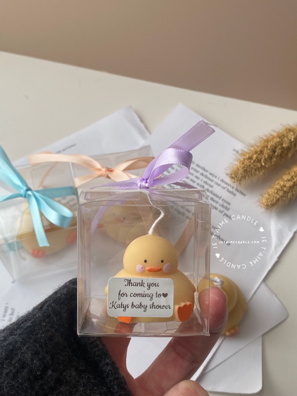 A close-up of four handcrafted duck-shaped candles in soft yellow tones with orange beaks and feet, and pink blushed cheeks, ideal for cute gifts or decorative accents. Three of the duck candles are packaged in clear plastic gift boxes tied with pastel-colored ribbon bows in blue, lavender, and peach, while one candle is displayed unboxed on a layered stack of white papers for a natural, cozy setting. The candles are labeled with 'Je T’aime Candle' tags and are placed beside a dried floral arrangement, creating a soft, aesthetic backdrop. These adorable soy wax candles are perfect for baby showers, birthday favors, Easter gifts, or aesthetic room decor.