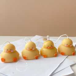 A close-up of four handcrafted duck-shaped candles in soft yellow tones with orange beaks and feet, and pink blushed cheeks, ideal for cute gifts or decorative accents. The candles are labeled with 'Je T’aime Candle' tags and are placed beside a dried floral arrangement, creating a soft, aesthetic backdrop. These adorable soy wax candles are perfect for baby showers, birthday favors, Easter gifts, or aesthetic room decor.