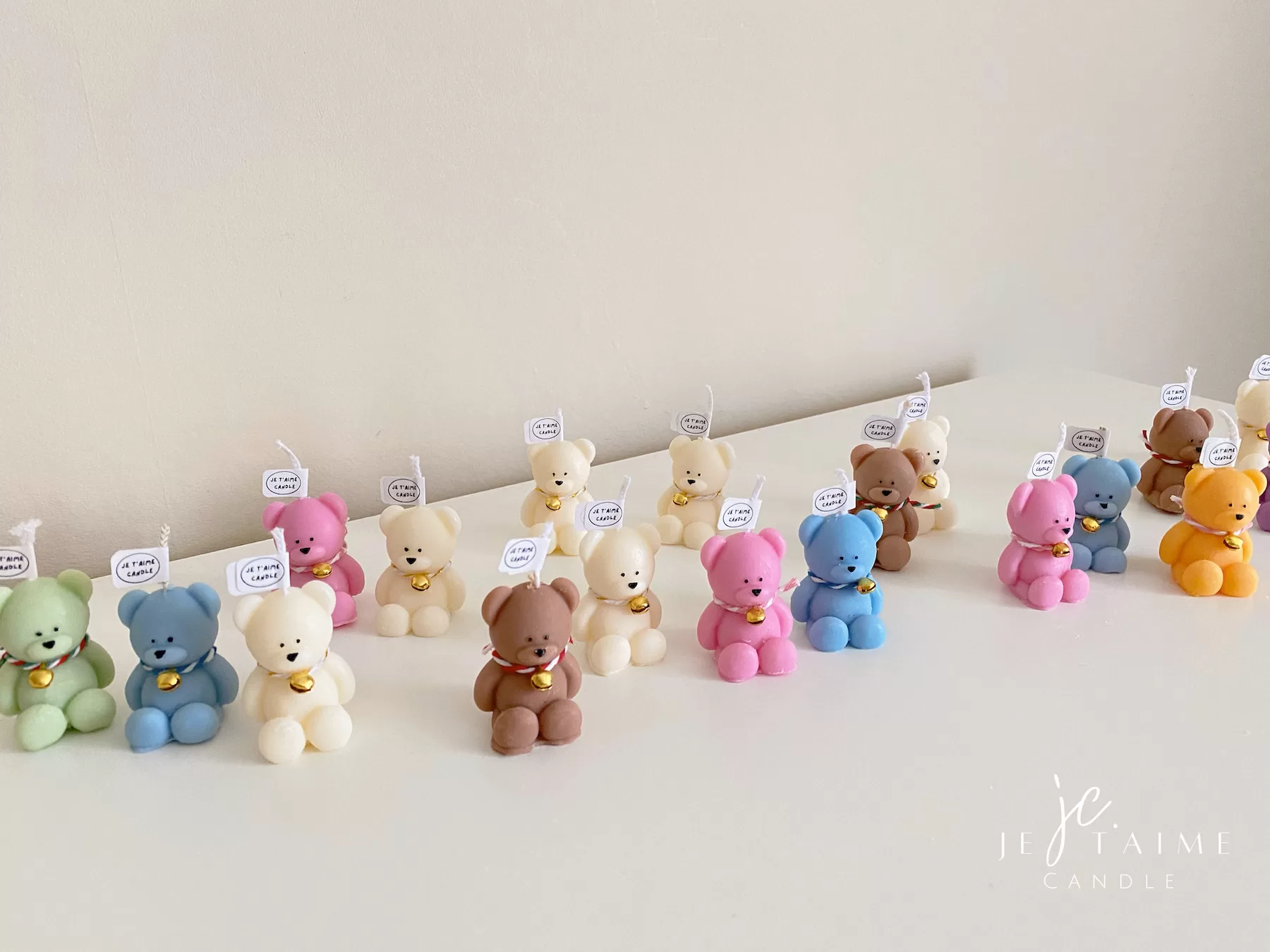 Colorful teddy bear candles in pink, blue, brown, orange, and white, wrapped in white organza bags with ribbons and personalized tags, displayed on a green tiled box with pampas grass decor in the background