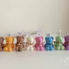 Colorful teddy bear candles in pink, green, blue, brown, orange, and white, wrapped in white organza bags with ribbons and personalized tags, displayed on a green tiled box with pampas grass decor in the background