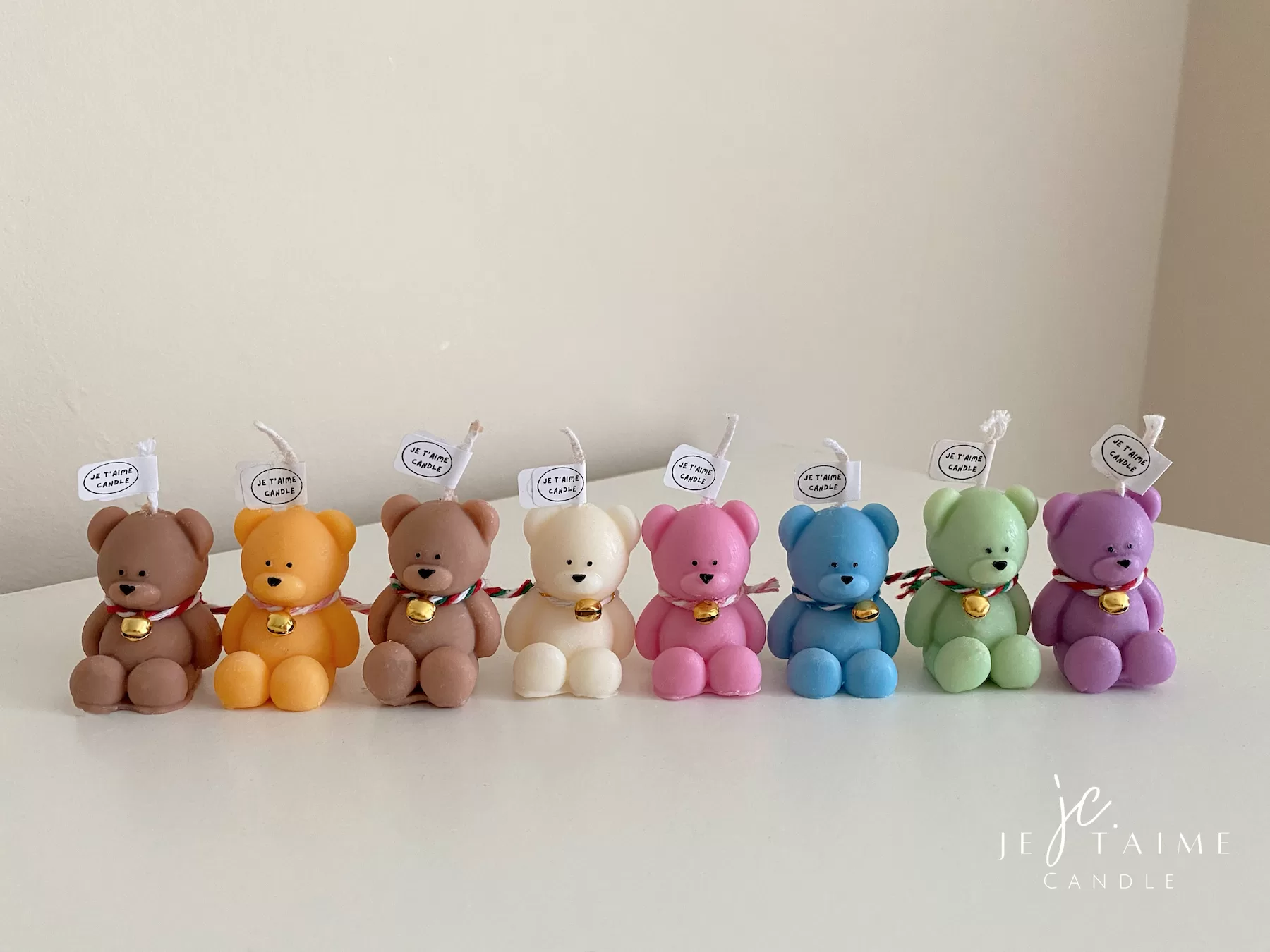 Colorful teddy bear candles in pink, green, blue, brown, orange, and white, wrapped in white organza bags with ribbons and personalized tags, displayed on a green tiled box with pampas grass decor in the background