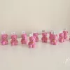 Colorful teddy bear candles in pink, wrapped in white organza bags with ribbons and personalized tags, displayed on a green tiled box with pampas grass decor in the background