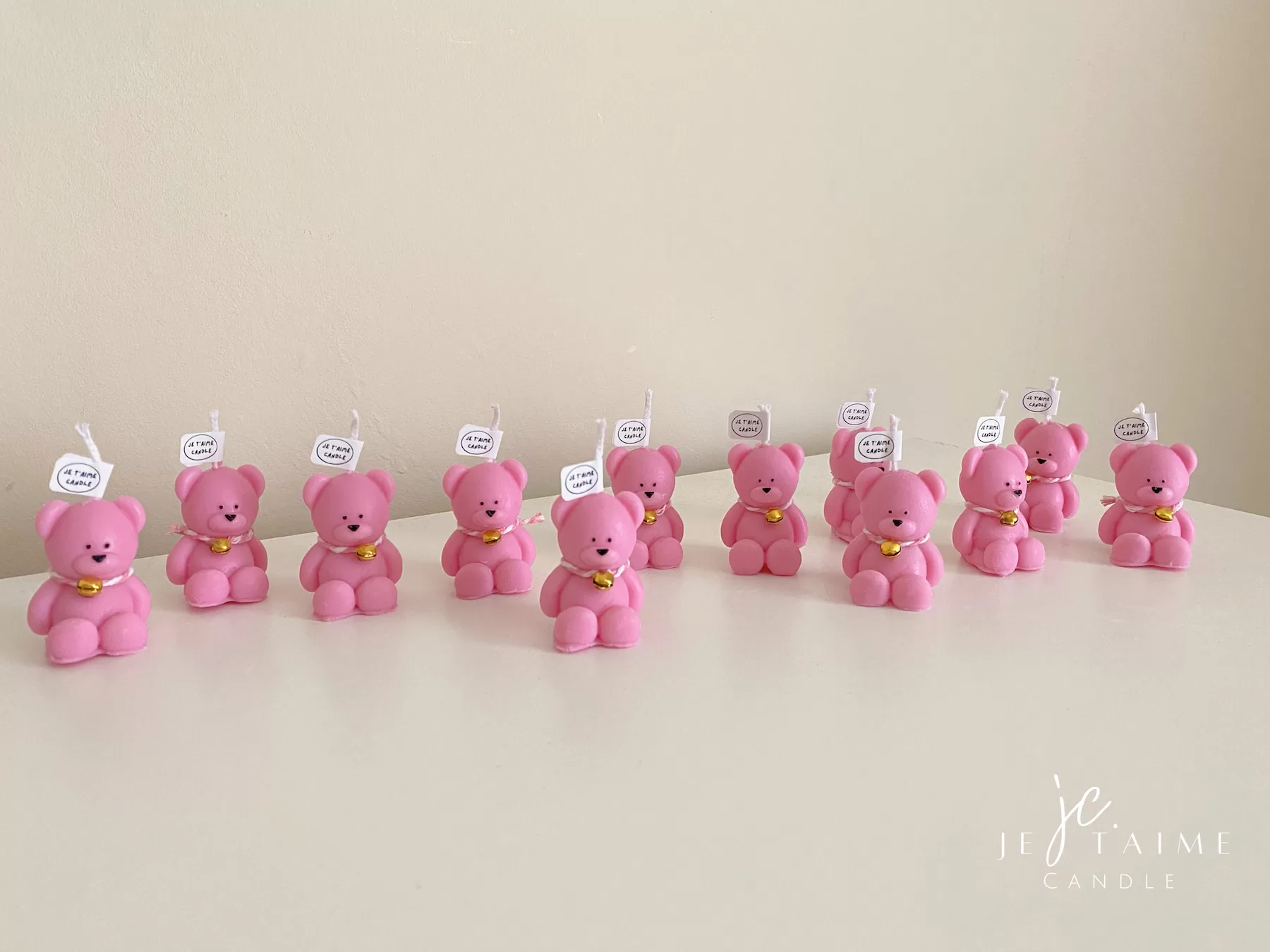 Colorful teddy bear candles in pink, wrapped in white organza bags with ribbons and personalized tags, displayed on a green tiled box with pampas grass decor in the background