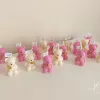 Colorful teddy bear candles in pink, white, wrapped in white organza bags with ribbons and personalized tags, displayed on a green tiled box with pampas grass decor in the background