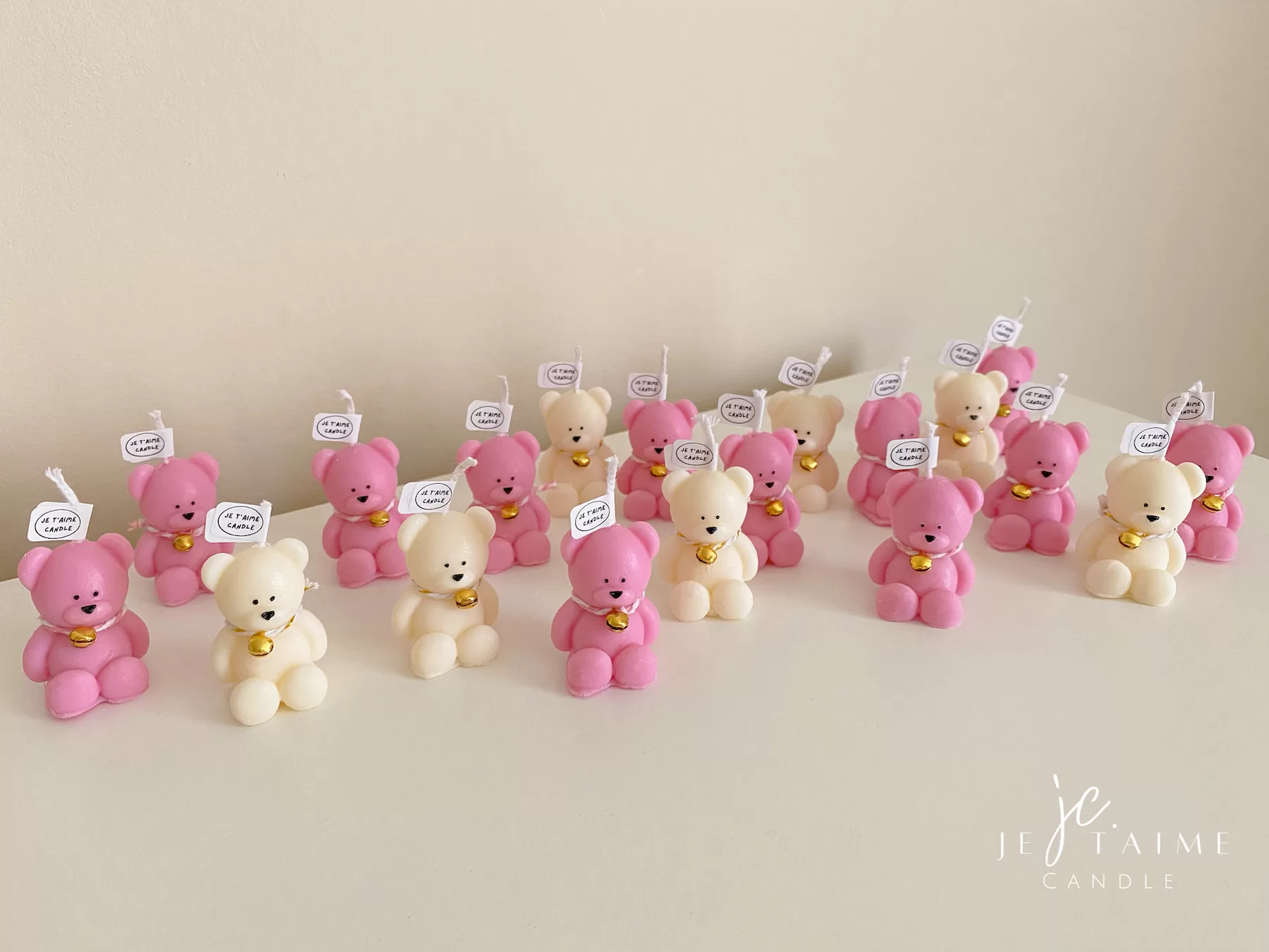 Colorful teddy bear candles in pink, white, wrapped in white organza bags with ribbons and personalized tags, displayed on a green tiled box with pampas grass decor in the background