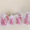 Colorful teddy bear candles in pink, white, wrapped in white organza bags with ribbons and personalized tags, displayed on a green tiled box with pampas grass decor in the background