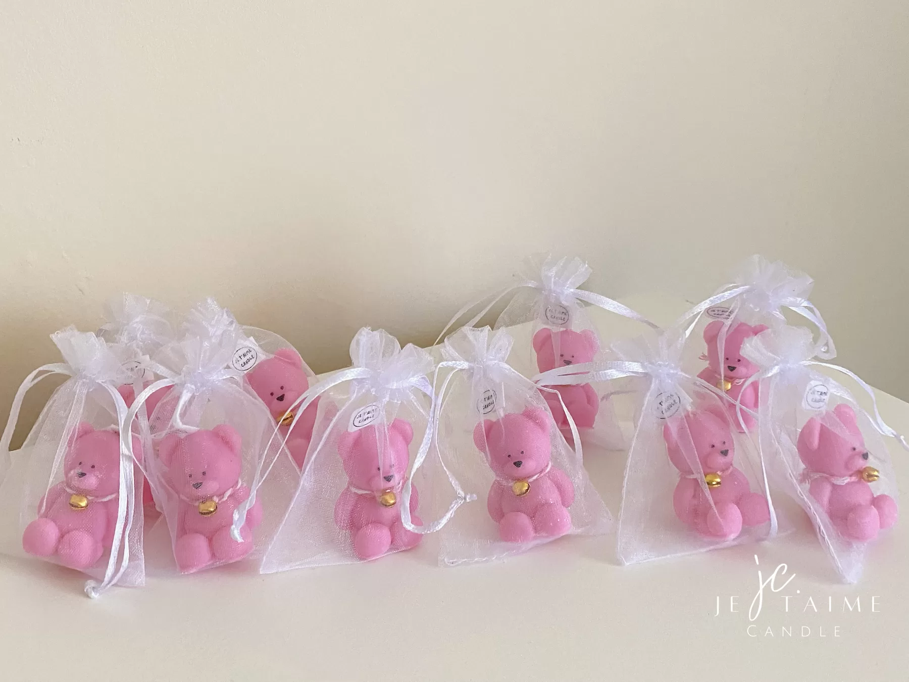 Colorful teddy bear candles in pink, white, wrapped in white organza bags with ribbons and personalized tags, displayed on a green tiled box with pampas grass decor in the background