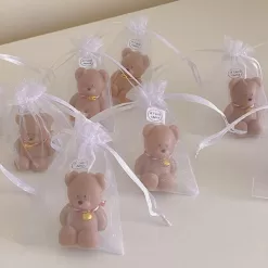 Colorful teddy bear candles in brown, wrapped in white organza bags with ribbons and personalized tags, displayed on a green tiled box with pampas grass decor in the background