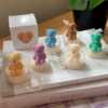 A collection of whimsical handmade bear-shaped candles in soft pastel colors, arranged on a cozy table. Each candle features intricate details. The candles are perfect for nursery decor, gifts, or home accents, adding warmth and charm to any space.