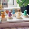 A collection of whimsical handmade bear-shaped candles in soft pastel colors, arranged on a cozy table. Each candle features intricate details. The candles are perfect for nursery decor, gifts, or home accents, adding warmth and charm to any space.