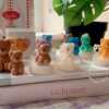 A collection of whimsical handmade bear-shaped candles in soft pastel colors, arranged on a cozy table. Each candle features intricate details. The candles are perfect for nursery decor, gifts, or home accents, adding warmth and charm to any space.