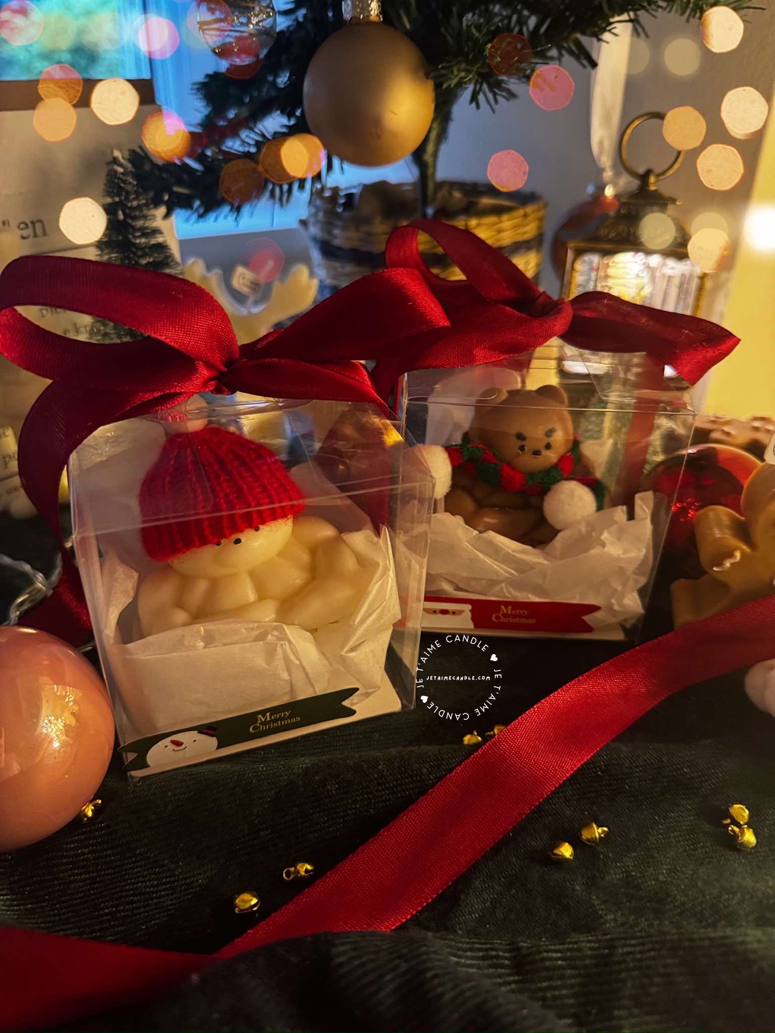 Two small bears on a table surrounded by festive Christmas decorations, one wearing a knitted hat and the other a scarf.