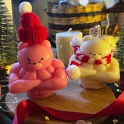 Two small bears on a table surrounded by festive Christmas decorations, one wearing a knitted hat and the other a scarf.