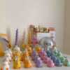 A colorful display of handmade duck-shaped candles in white, yellow, orange, lavender, and pink hues, arranged on a tabletop with charming pastel decor in the background. Each ducky candle features hand-painted blush cheeks, orange beaks, and feet, with labels reading 'JE T’AIME CANDLE'. Behind the duck candles, there are uniquely shaped spiral and flower candles in yellow and lavender, along with a pink bear candle, a bouquet of pink tulips, a mirrored disco mushroom lamp, and a white paper bag with French text used as part of the aesthetic room decor. This cute candle collection is ideal for baby shower gifts, birthday favors, desk decoration, or aesthetic home styling.