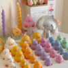 A colorful display of handmade duck-shaped candles in white, yellow, orange, lavender, and pink hues, arranged on a tabletop with charming pastel decor in the background. Each ducky candle features hand-painted blush cheeks, orange beaks, and feet, with labels reading 'JE T’AIME CANDLE'. Behind the duck candles, there are uniquely shaped spiral and flower candles in yellow and lavender, along with a pink bear candle, a bouquet of pink tulips, a mirrored disco mushroom lamp, and a white paper bag with French text used as part of the aesthetic room decor. This cute candle collection is ideal for baby shower gifts, birthday favors, desk decoration, or aesthetic home styling.