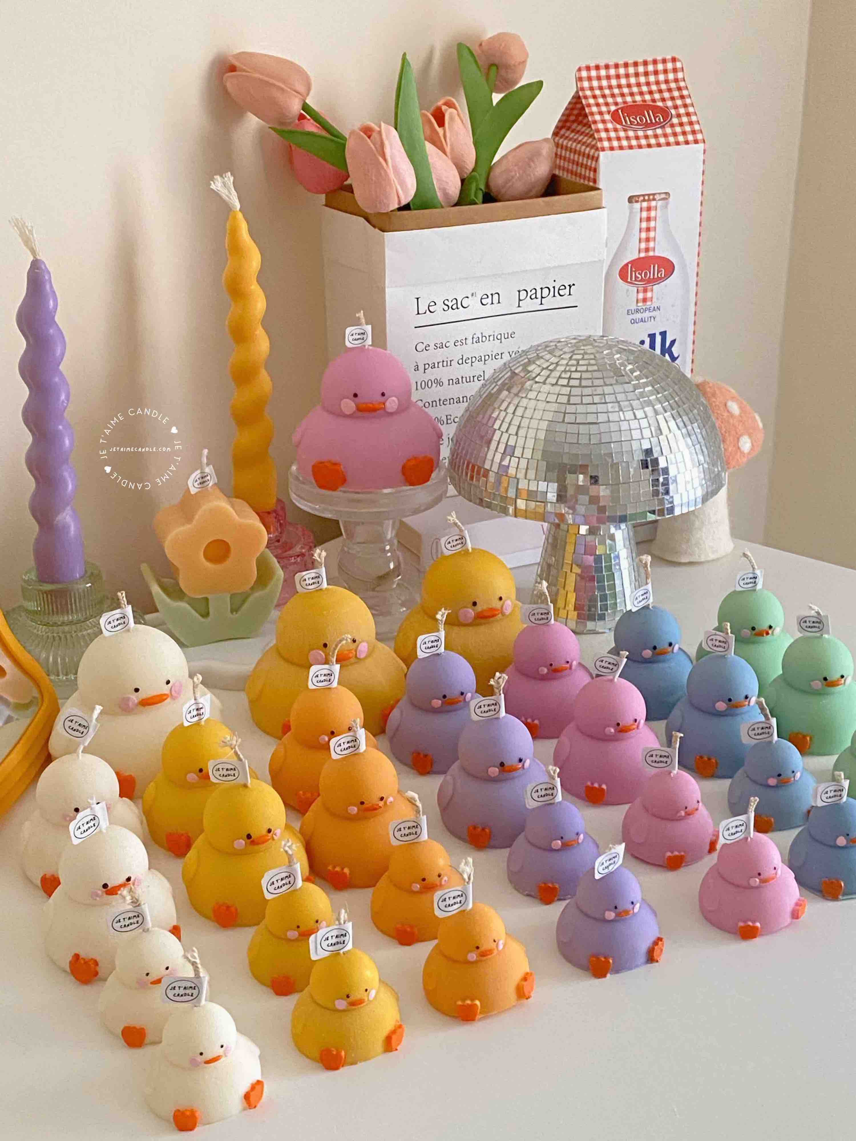 A colorful display of handmade duck-shaped candles in white, yellow, orange, lavender, and pink hues, arranged on a tabletop with charming pastel decor in the background. Each ducky candle features hand-painted blush cheeks, orange beaks, and feet, with labels reading 'JE T’AIME CANDLE'. Behind the duck candles, there are uniquely shaped spiral and flower candles in yellow and lavender, along with a pink bear candle, a bouquet of pink tulips, a mirrored disco mushroom lamp, and a white paper bag with French text used as part of the aesthetic room decor. This cute candle collection is ideal for baby shower gifts, birthday favors, desk decoration, or aesthetic home styling.