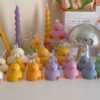 A colorful display of handmade duck-shaped candles in white, yellow, orange, lavender, and pink hues, arranged on a tabletop with charming pastel decor in the background. Each ducky candle features hand-painted blush cheeks, orange beaks, and feet, with labels reading 'JE T’AIME CANDLE'. Behind the duck candles, there are uniquely shaped spiral and flower candles in yellow and lavender, along with a pink bear candle, a bouquet of pink tulips, a mirrored disco mushroom lamp, and a white paper bag with French text used as part of the aesthetic room decor. This cute candle collection is ideal for baby shower gifts, birthday favors, desk decoration, or aesthetic home styling.