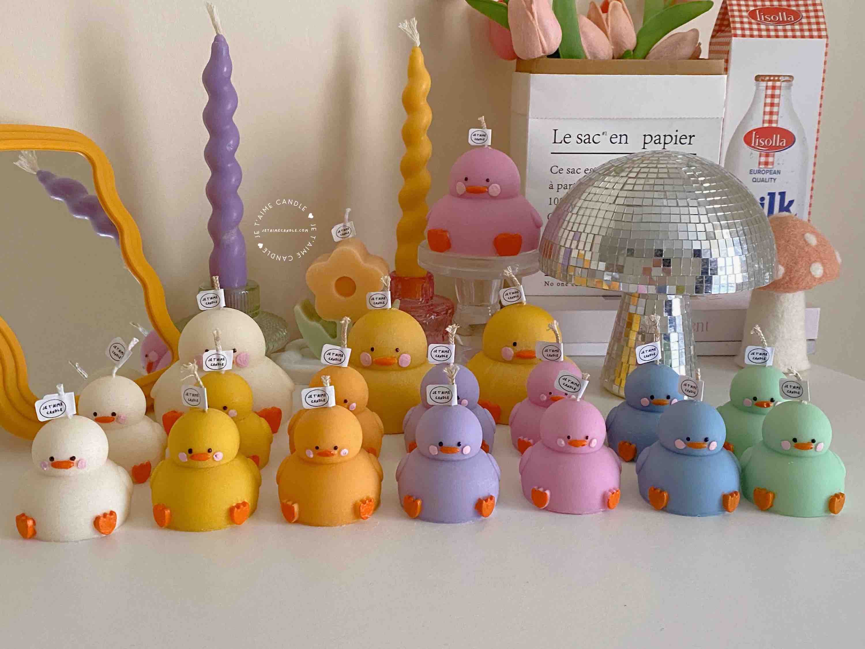 A colorful display of handmade duck-shaped candles in white, yellow, orange, lavender, and pink hues, arranged on a tabletop with charming pastel decor in the background. Each ducky candle features hand-painted blush cheeks, orange beaks, and feet, with labels reading 'JE T’AIME CANDLE'. Behind the duck candles, there are uniquely shaped spiral and flower candles in yellow and lavender, along with a pink bear candle, a bouquet of pink tulips, a mirrored disco mushroom lamp, and a white paper bag with French text used as part of the aesthetic room decor. This cute candle collection is ideal for baby shower gifts, birthday favors, desk decoration, or aesthetic home styling.
