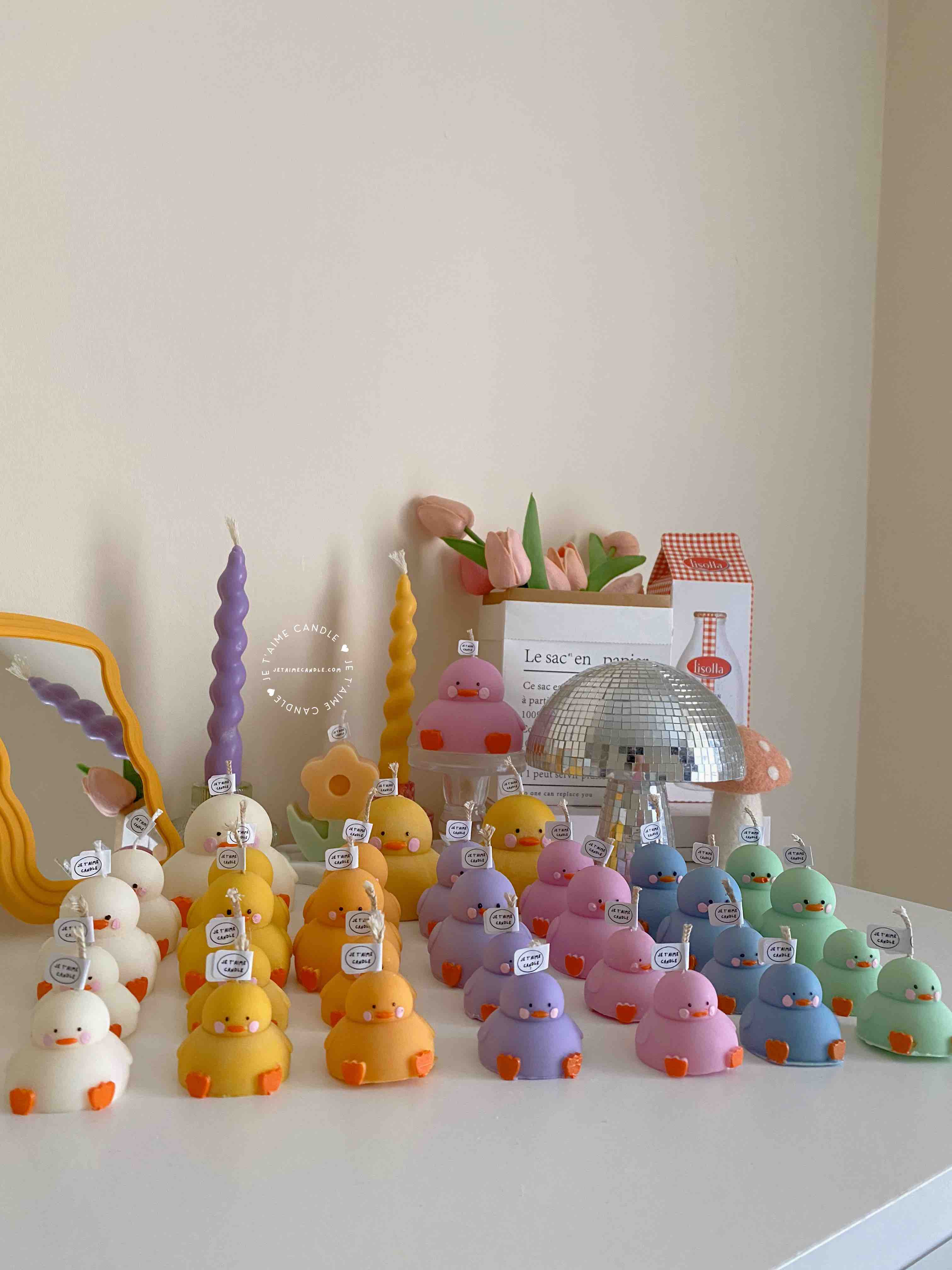 A colorful display of handmade duck-shaped candles in white, yellow, orange, lavender, and pink hues, arranged on a tabletop with charming pastel decor in the background. Each ducky candle features hand-painted blush cheeks, orange beaks, and feet, with labels reading 'JE T’AIME CANDLE'. Behind the duck candles, there are uniquely shaped spiral and flower candles in yellow and lavender, along with a pink bear candle, a bouquet of pink tulips, a mirrored disco mushroom lamp, and a white paper bag with French text used as part of the aesthetic room decor. This cute candle collection is ideal for baby shower gifts, birthday favors, desk decoration, or aesthetic home styling.