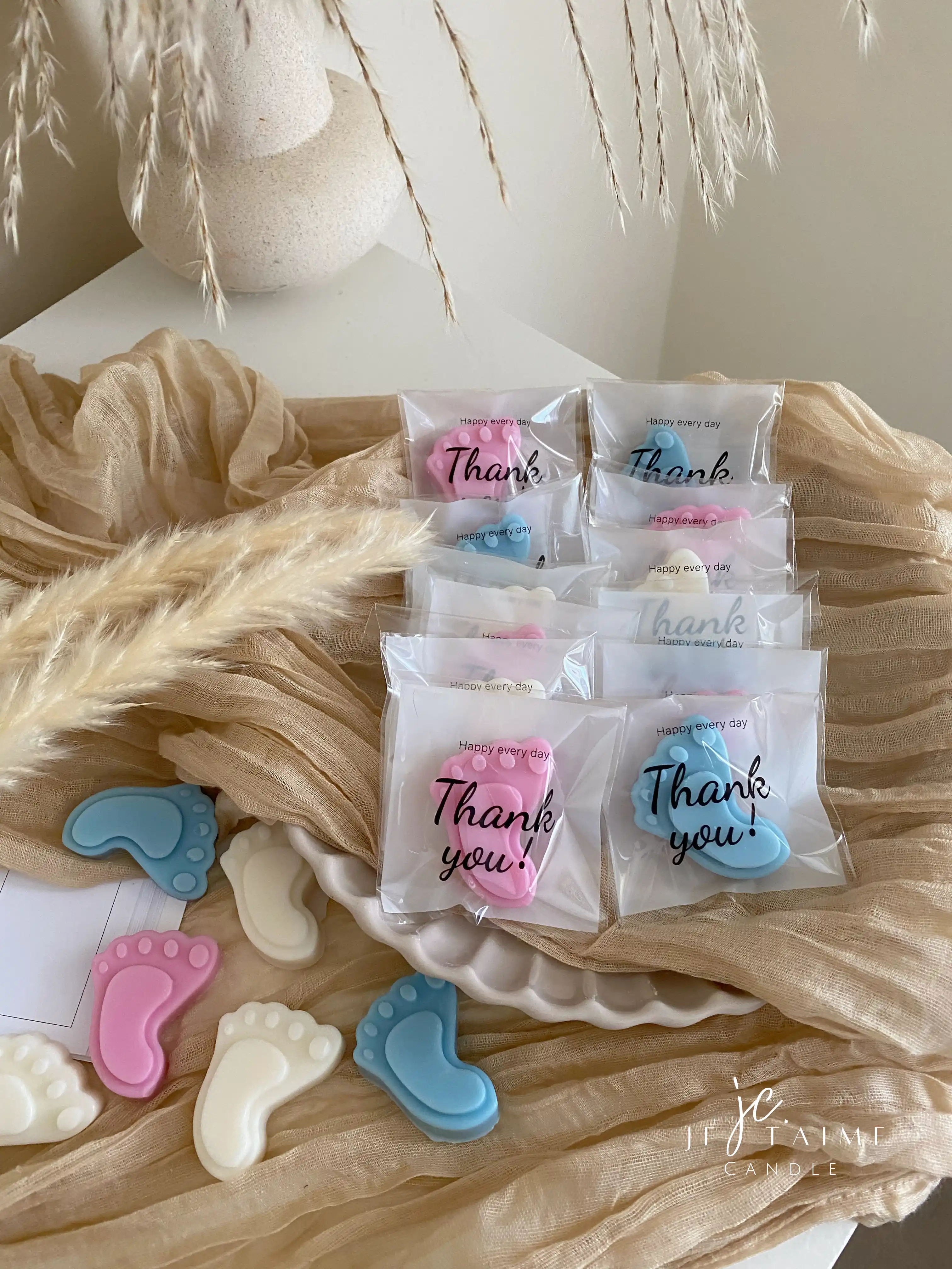This image showcases beautifully packaged baby shower favors in the shape of small baby feet, made of wax. The favors come in pastel colors, including pink, blue, and white, and are individually wrapped in clear plastic with a "Thank you!" message on each package. The elegant presentation, with soft fabric, pampas grass, and ceramic dishware, creates a warm and delicate aesthetic. These favors are perfect for baby showers, gender reveals, or newborn celebrations.