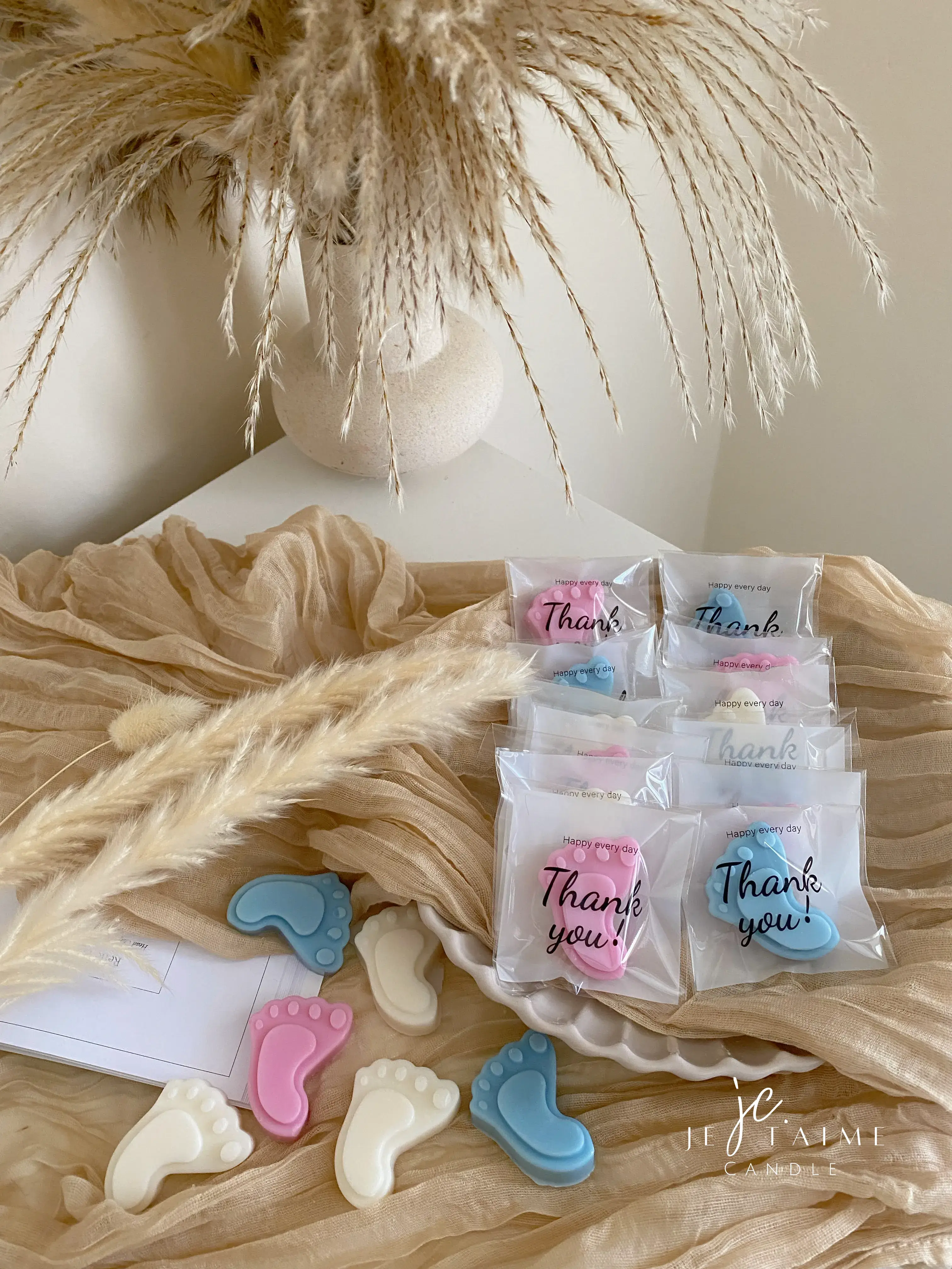 This image showcases beautifully packaged baby shower favors in the shape of small baby feet, made of wax. The favors come in pastel colors, including pink, blue, and white, and are individually wrapped in clear plastic with a "Thank you!" message on each package. The elegant presentation, with soft fabric, pampas grass, and ceramic dishware, creates a warm and delicate aesthetic. These favors are perfect for baby showers, gender reveals, or newborn celebrations.