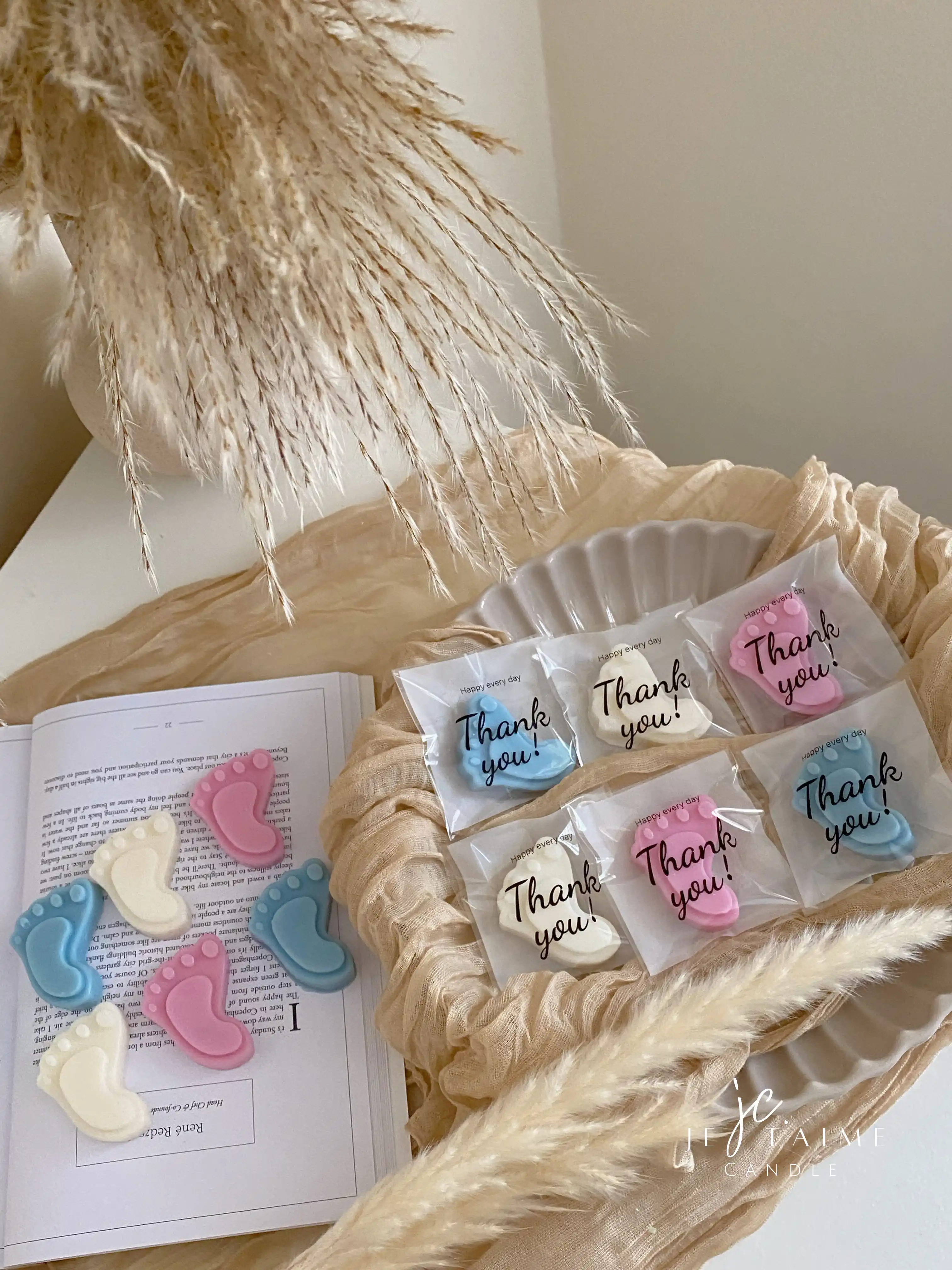 This image showcases beautifully packaged baby shower favors in the shape of small baby feet, made of wax. The favors come in pastel colors, including pink, blue, and white, and are individually wrapped in clear plastic with a "Thank you!" message on each package. The elegant presentation, with soft fabric, pampas grass, and ceramic dishware, creates a warm and delicate aesthetic. These favors are perfect for baby showers, gender reveals, or newborn celebrations.