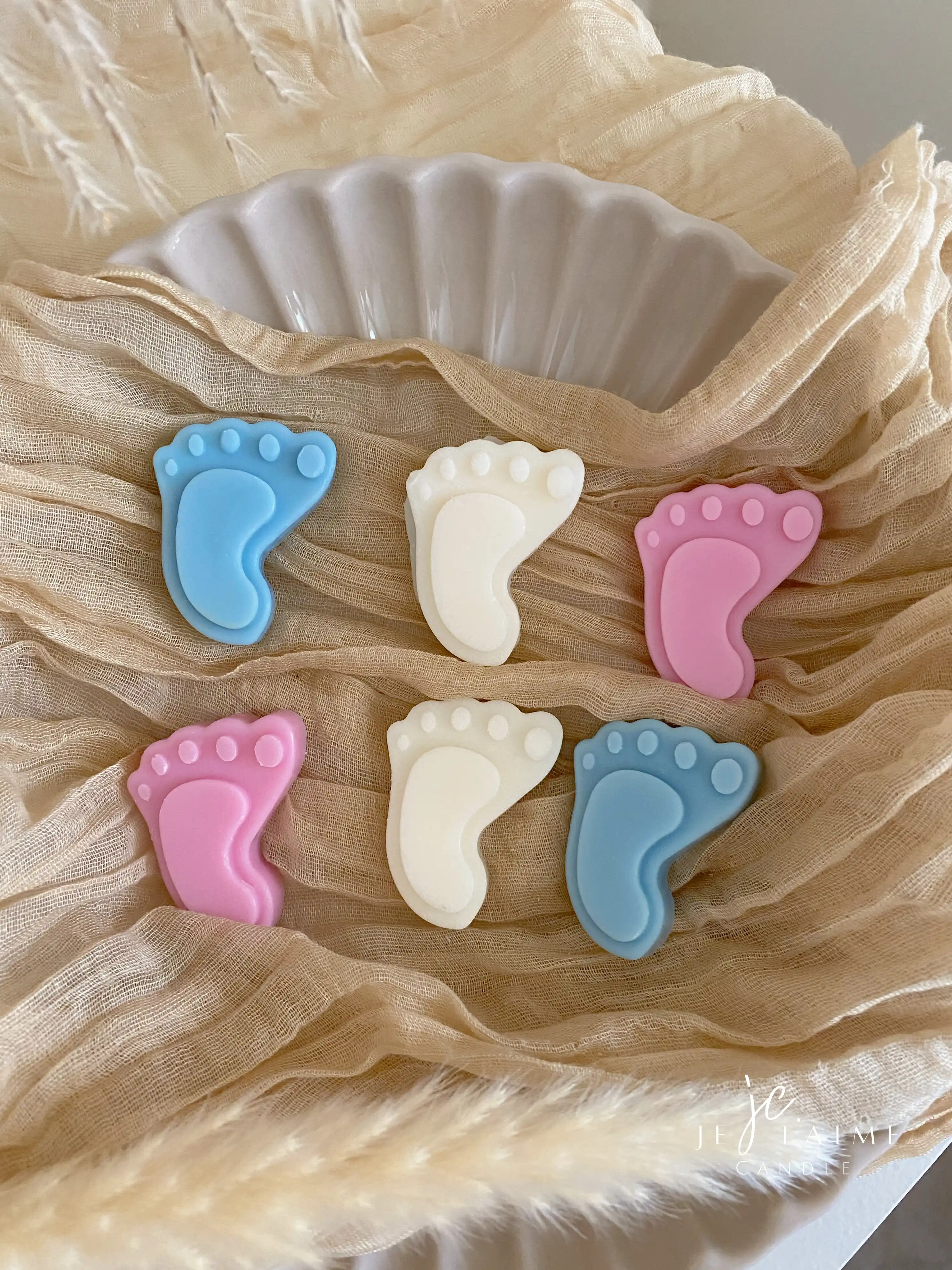 This image showcases beautifully packaged baby shower favors in the shape of small baby feet, made of wax. The favors come in pastel colors, including pink, blue, and white, and are individually wrapped in clear plastic with a "Thank you!" message on each package. The elegant presentation, with soft fabric, pampas grass, and ceramic dishware, creates a warm and delicate aesthetic. These favors are perfect for baby showers, gender reveals, or newborn celebrations.