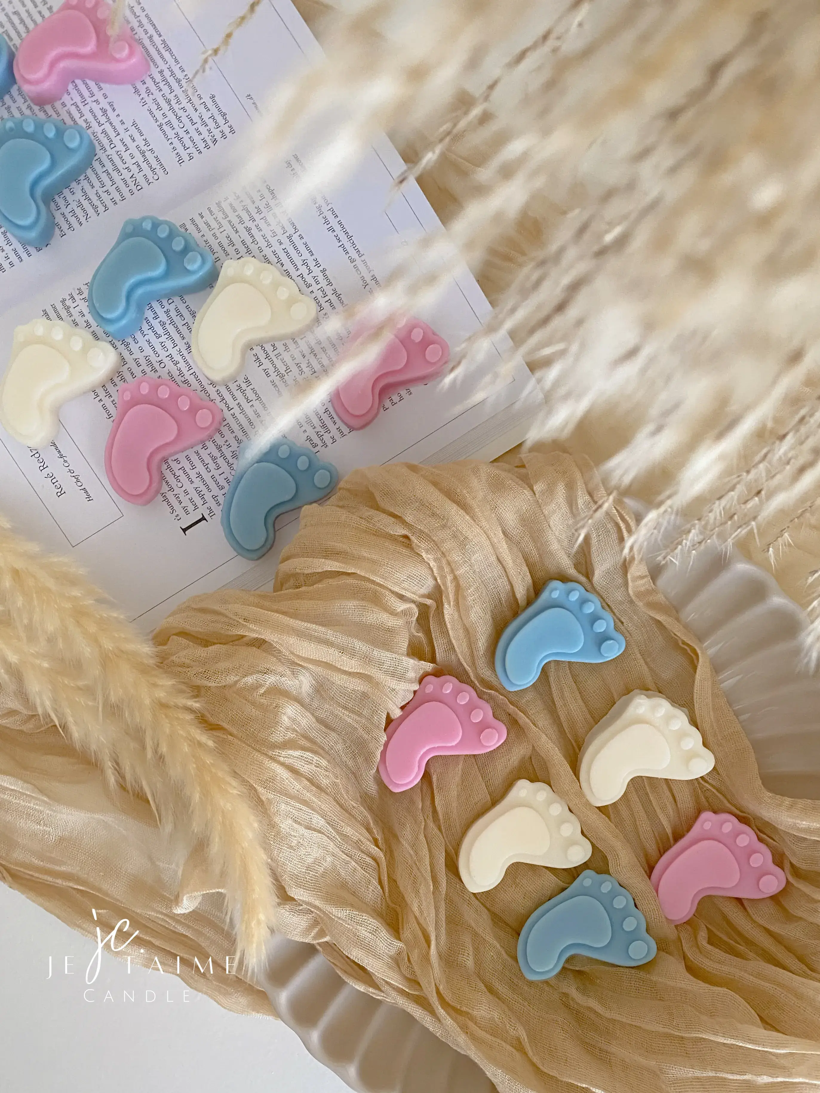 This image showcases beautifully packaged baby shower favors in the shape of small baby feet, made of wax. The favors come in pastel colors, including pink, blue, and white, and are individually wrapped in clear plastic with a "Thank you!" message on each package. The elegant presentation, with soft fabric, pampas grass, and ceramic dishware, creates a warm and delicate aesthetic. These favors are perfect for baby showers, gender reveals, or newborn celebrations.