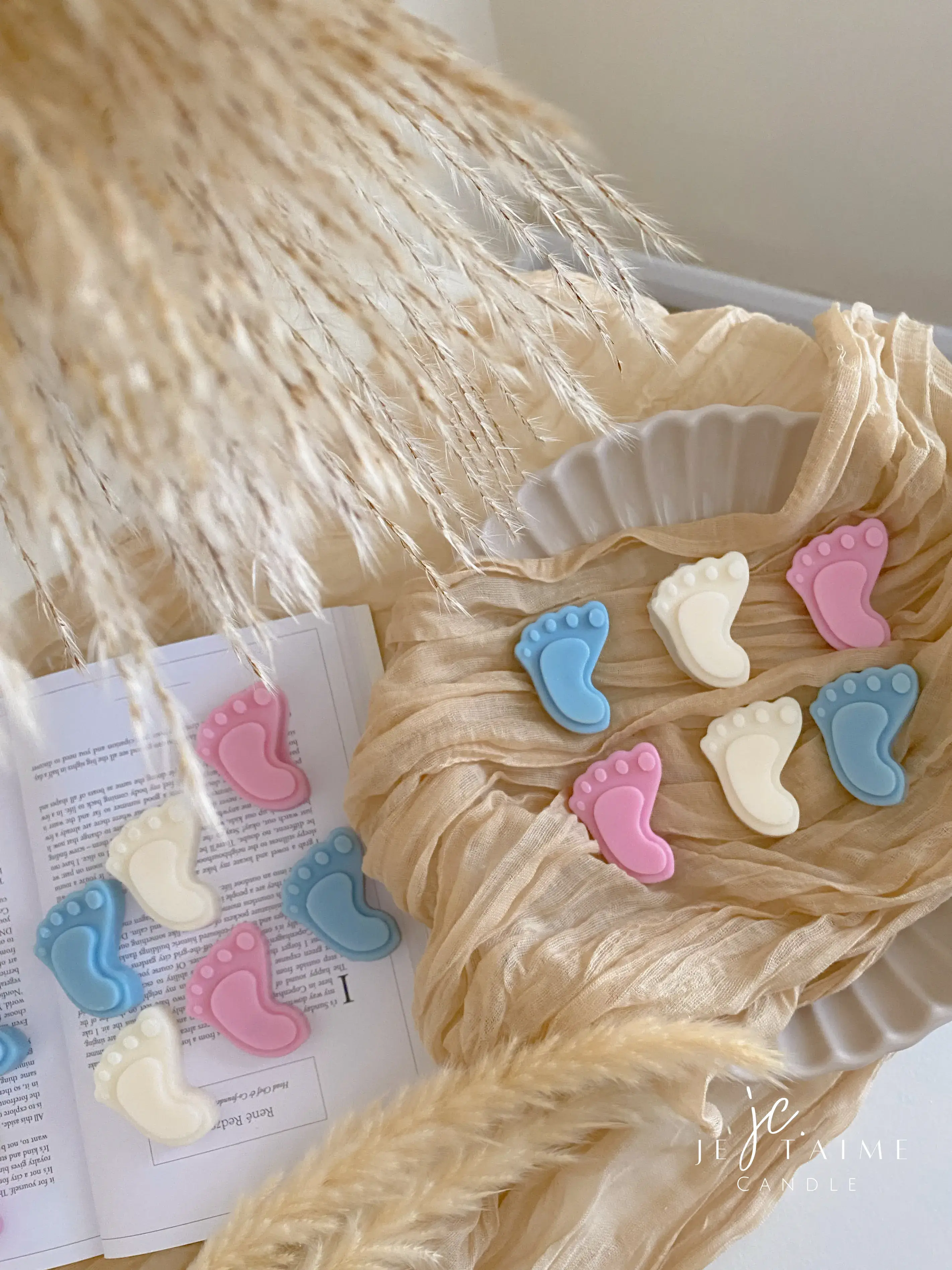 This image showcases beautifully packaged baby shower favors in the shape of small baby feet, made of wax. The favors come in pastel colors, including pink, blue, and white, and are individually wrapped in clear plastic with a "Thank you!" message on each package. The elegant presentation, with soft fabric, pampas grass, and ceramic dishware, creates a warm and delicate aesthetic. These favors are perfect for baby showers, gender reveals, or newborn celebrations.