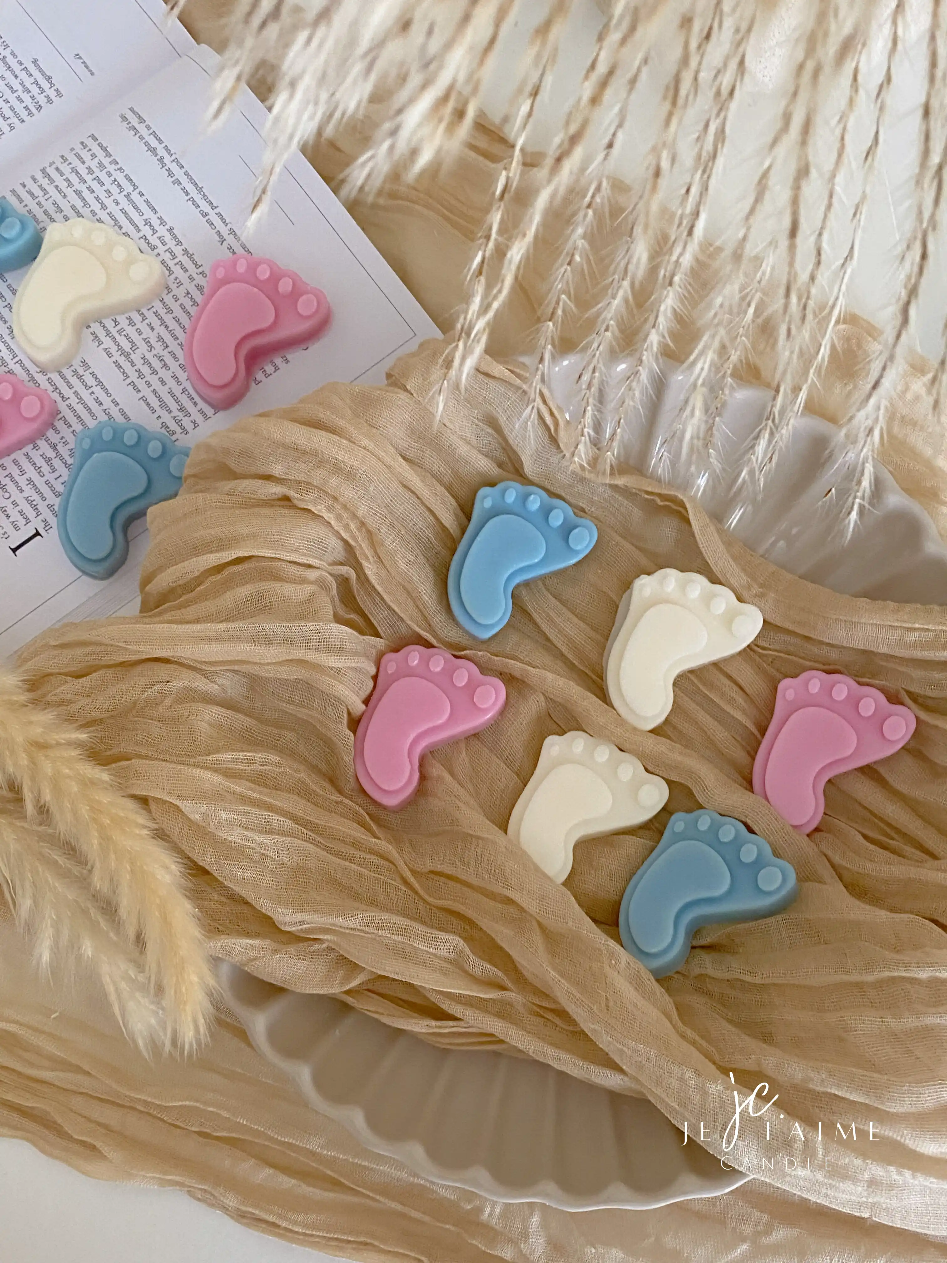 This image showcases beautifully packaged baby shower favors in the shape of small baby feet, made of wax. The favors come in pastel colors, including pink, blue, and white, and are individually wrapped in clear plastic with a "Thank you!" message on each package. The elegant presentation, with soft fabric, pampas grass, and ceramic dishware, creates a warm and delicate aesthetic. These favors are perfect for baby showers, gender reveals, or newborn celebrations.