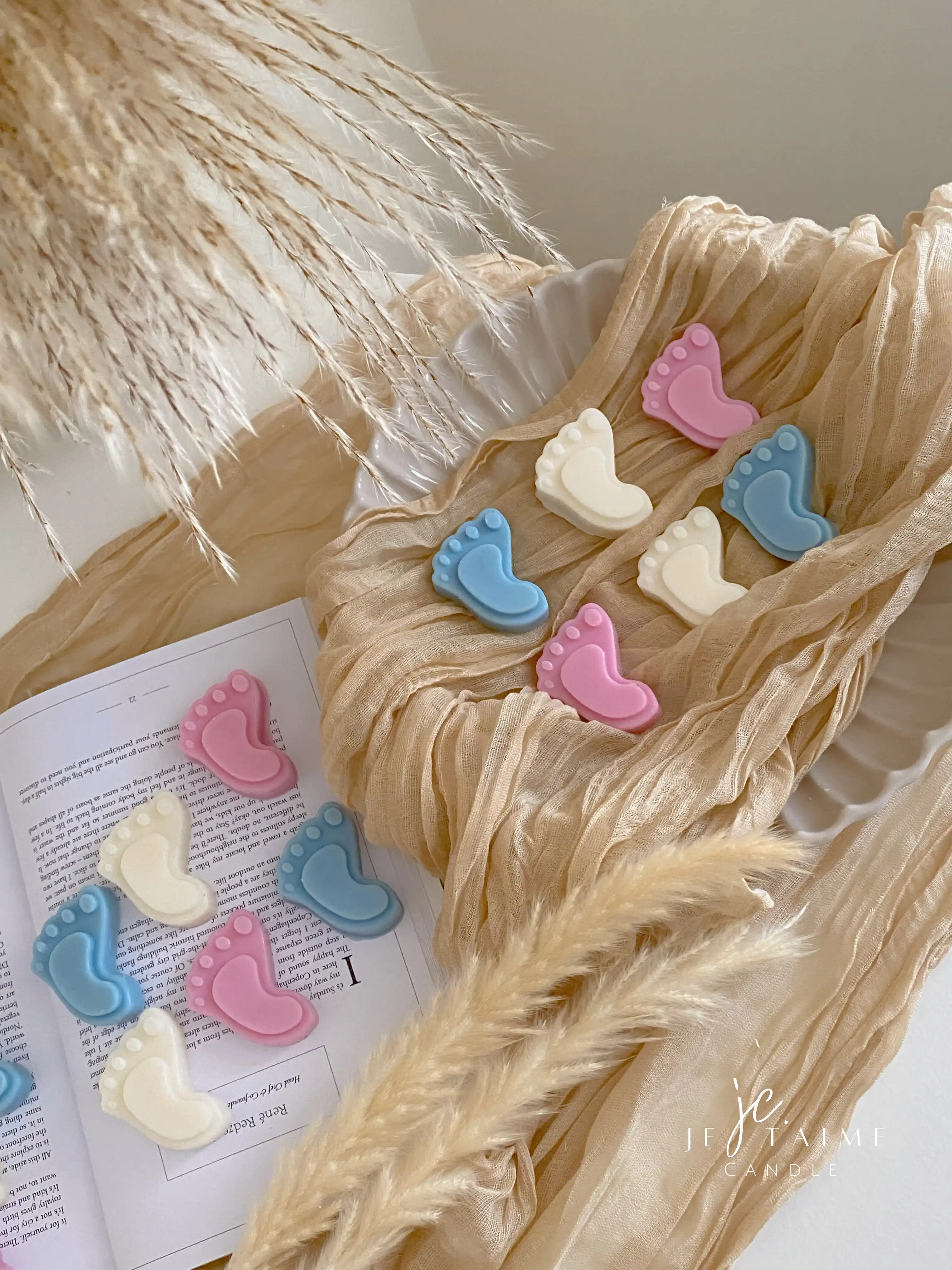 This image showcases beautifully packaged baby shower favors in the shape of small baby feet, made of wax. The favors come in pastel colors, including pink, blue, and white, and are individually wrapped in clear plastic with a "Thank you!" message on each package. The elegant presentation, with soft fabric, pampas grass, and ceramic dishware, creates a warm and delicate aesthetic. These favors are perfect for baby showers, gender reveals, or newborn celebrations.