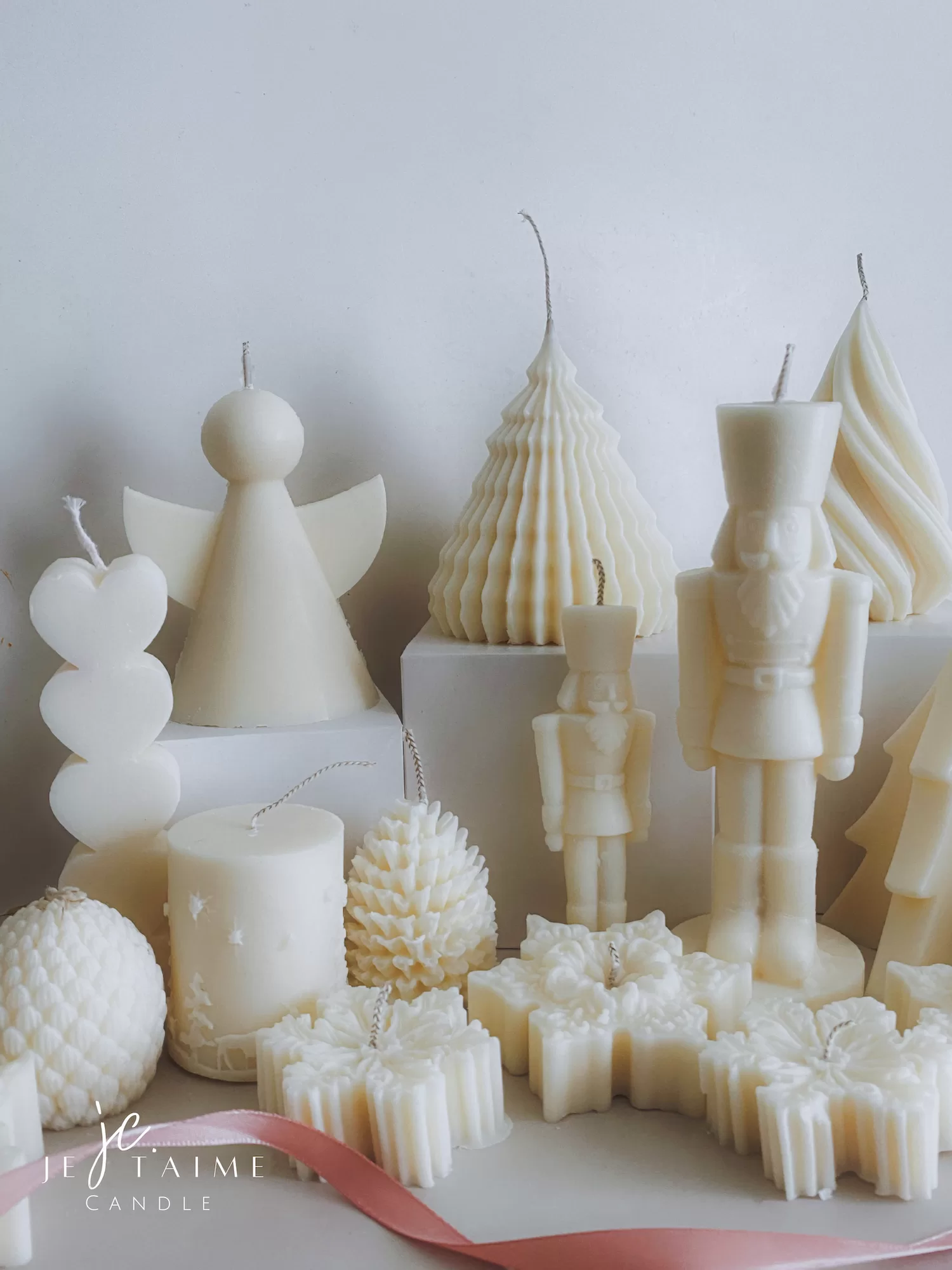 ollection of handmade decorative candles in Christmas shapes including houses, nutcrackers, snowflakes, pine cones, and Christmas trees, displayed on soft fabric with a pink ribbon.
