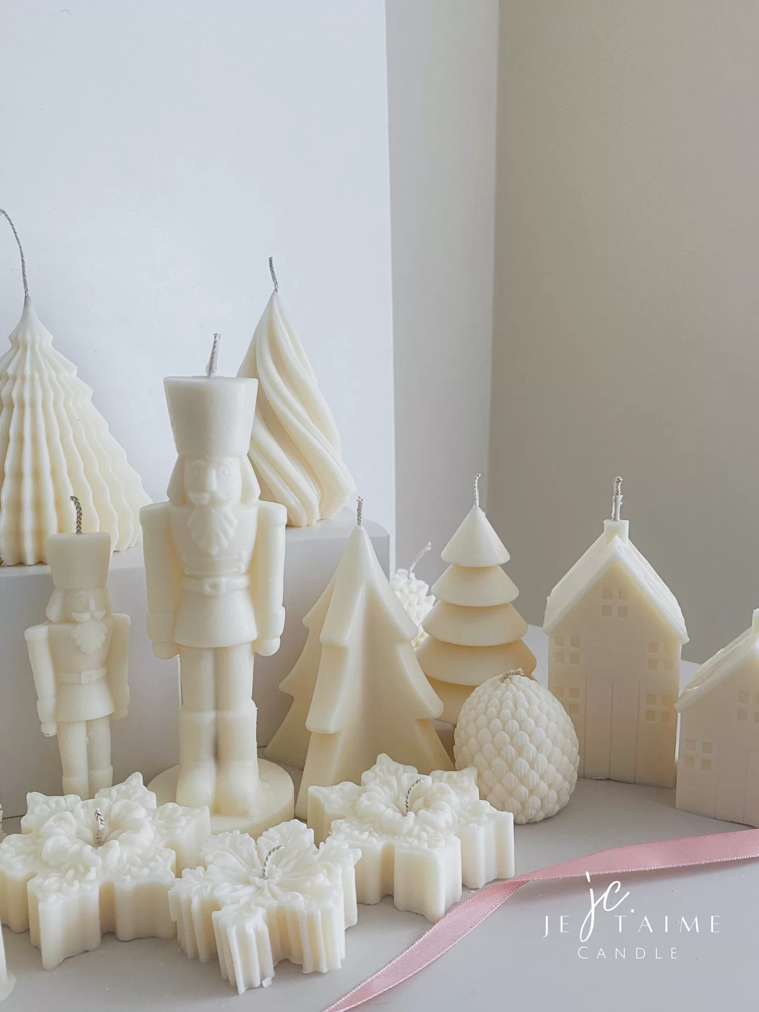 ollection of handmade decorative candles in Christmas shapes including houses, nutcrackers, snowflakes, pine cones, and Christmas trees, displayed on soft fabric with a pink ribbon.