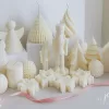 ollection of handmade decorative candles in Christmas shapes including houses, nutcrackers, snowflakes, pine cones, and Christmas trees, displayed on soft fabric with a pink ribbon.