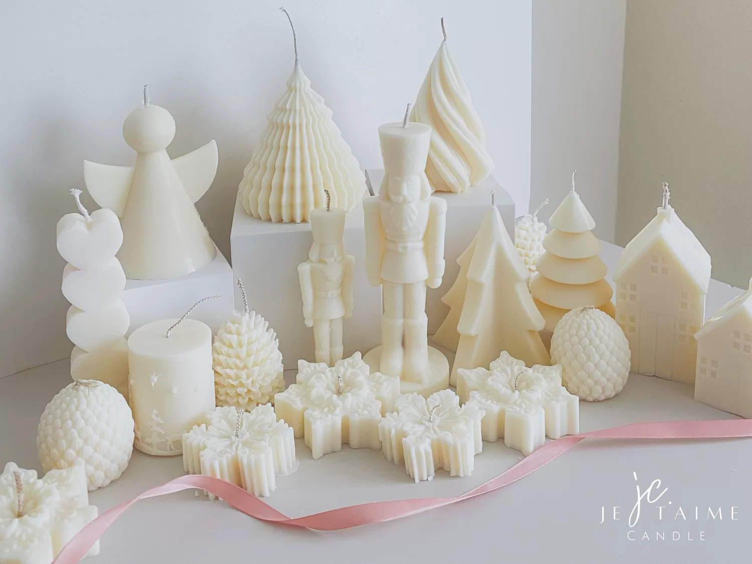 ollection of handmade decorative candles in Christmas shapes including houses, nutcrackers, snowflakes, pine cones, and Christmas trees, displayed on soft fabric with a pink ribbon.