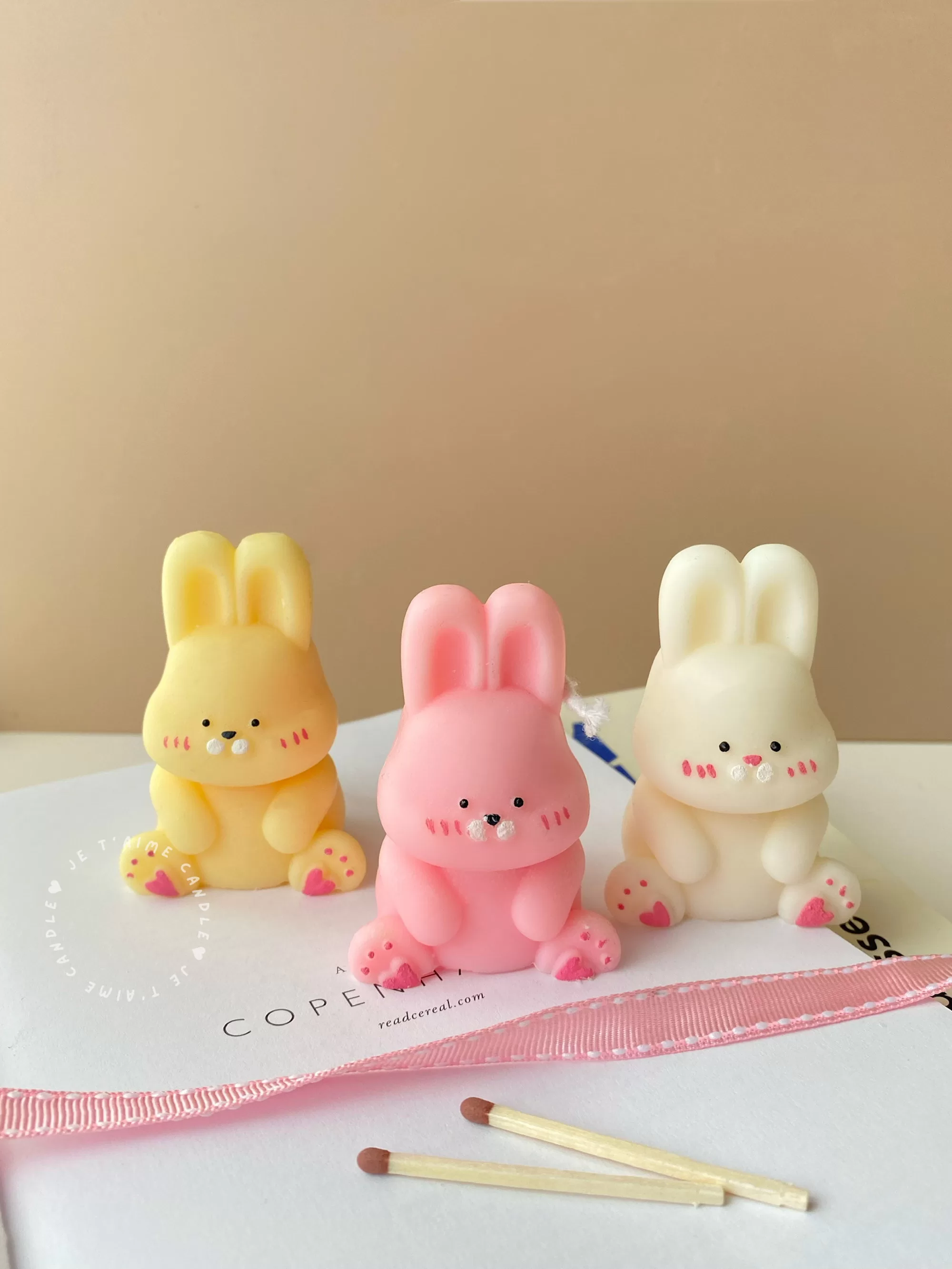 Three cute chubby bunny-shaped candles in pastel colors—yellow, pink, and white—sitting on a white surface with pink ribbon and wooden matches. Each bunny candle features detailed facial expressions, rosy cheeks, and tiny paw prints with heart designs. These kawaii bunny candles are perfect for Easter décor, baby showers, birthday gifts, or cozy home decoration.