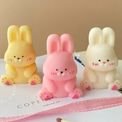 Three cute chubby bunny-shaped candles in pastel colors—yellow, pink, and white—sitting on a white surface with pink ribbon and wooden matches. Each bunny candle features detailed facial expressions, rosy cheeks, and tiny paw prints with heart designs. These kawaii bunny candles are perfect for Easter décor, baby showers, birthday gifts, or cozy home decoration.