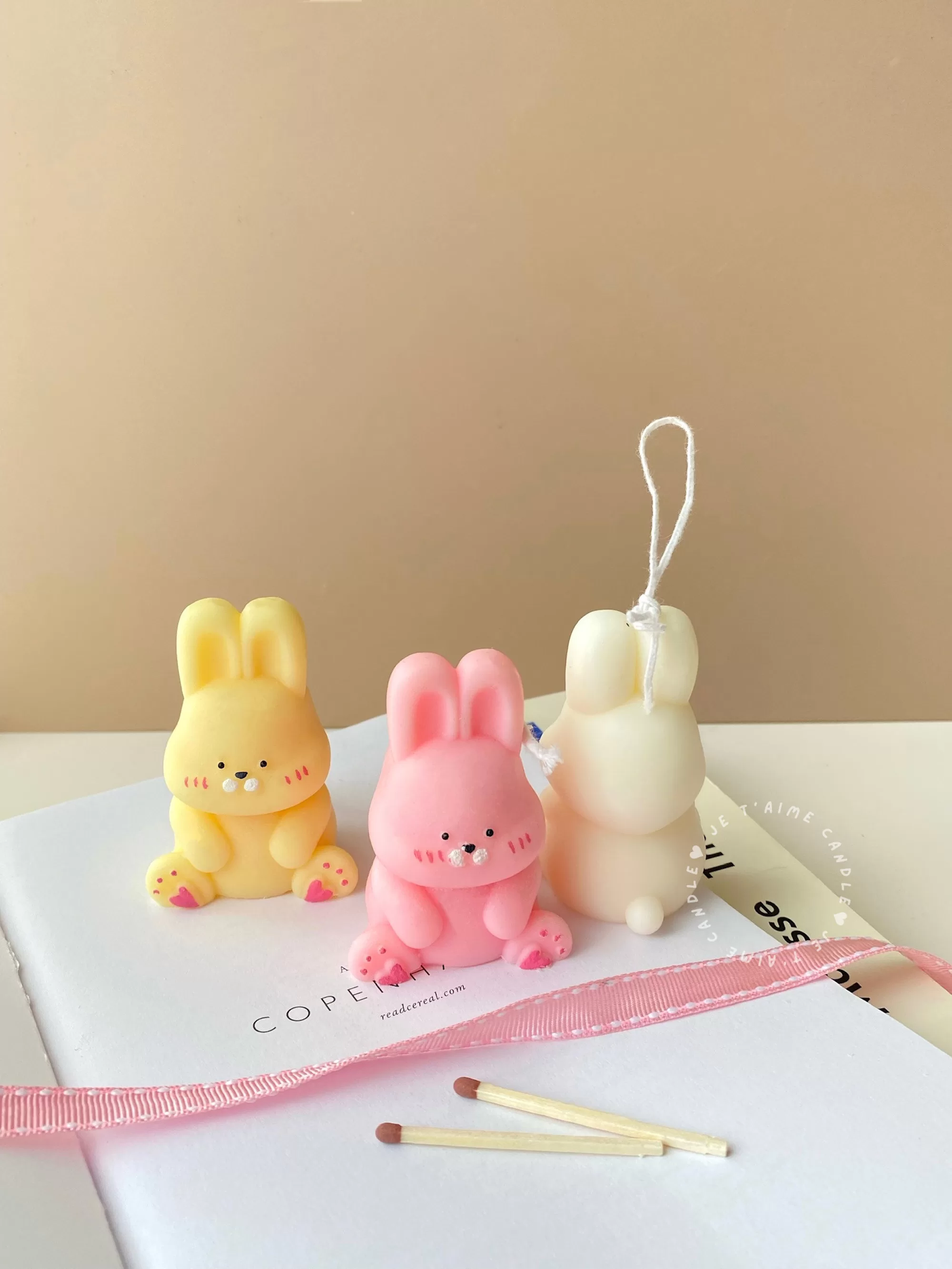 Three cute chubby bunny-shaped candles in pastel colors—yellow, pink, and white—sitting on a white surface with pink ribbon and wooden matches. Each bunny candle features detailed facial expressions, rosy cheeks, and tiny paw prints with heart designs. These kawaii bunny candles are perfect for Easter décor, baby showers, birthday gifts, or cozy home decoration.