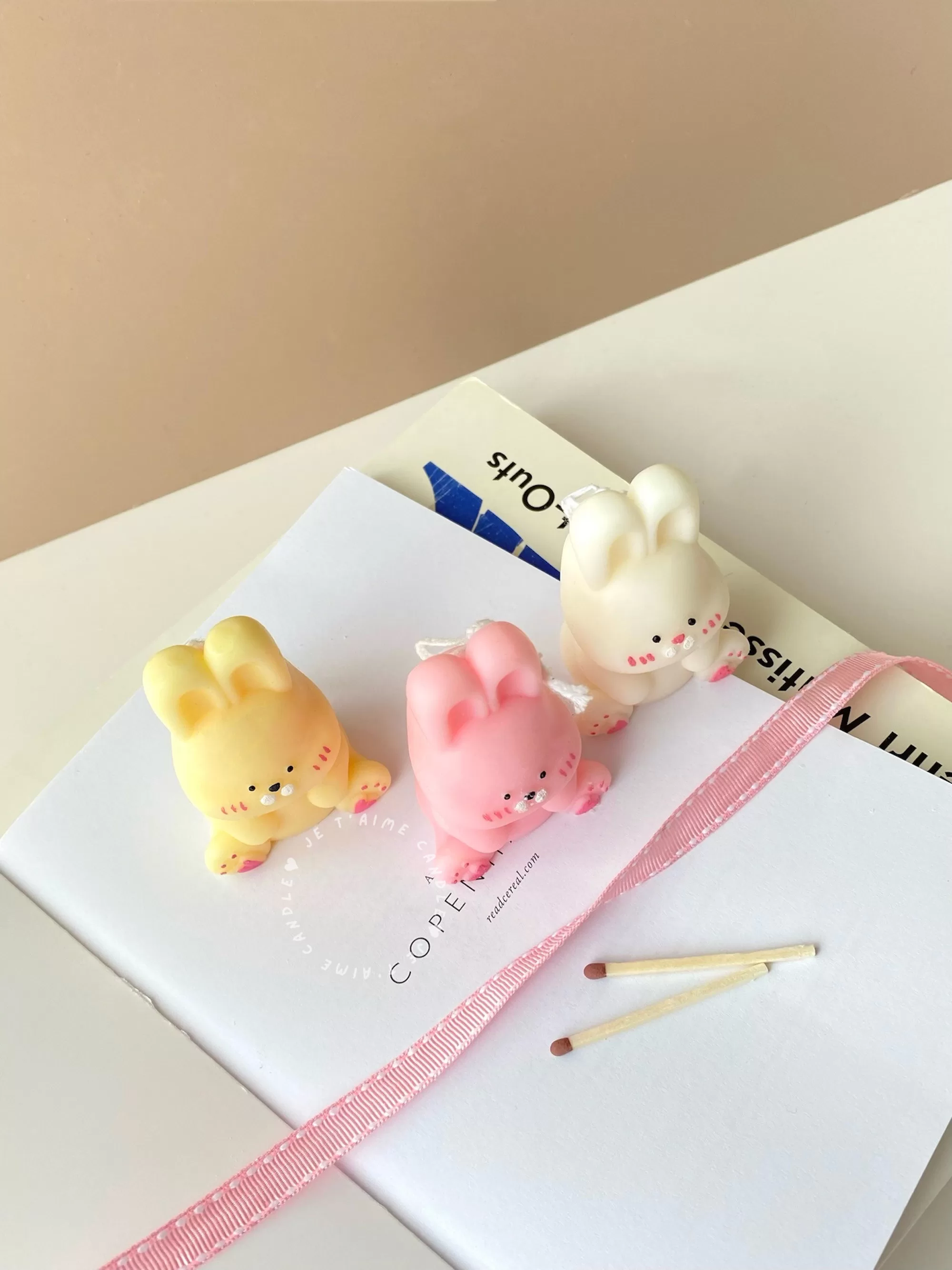 Three cute chubby bunny-shaped candles in pastel colors—yellow, pink, and white—sitting on a white surface with pink ribbon and wooden matches. Each bunny candle features detailed facial expressions, rosy cheeks, and tiny paw prints with heart designs. These kawaii bunny candles are perfect for Easter décor, baby showers, birthday gifts, or cozy home decoration.